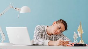Teenager with eyeglasses contemplating medals at desk with laptop and trophies.